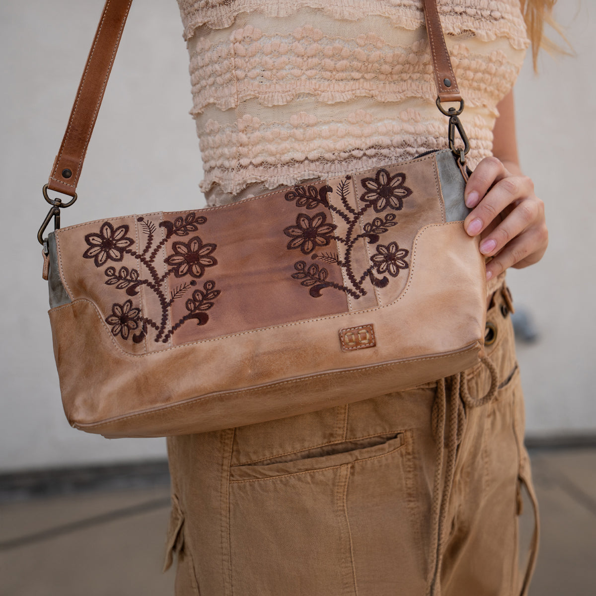 A person in a textured beige top and tan pants holds the Bed Stu Hyperbolic, a handcrafted leather shoulder bag with floral embroidery and a brown strap, adding a chic, bohemian touch.