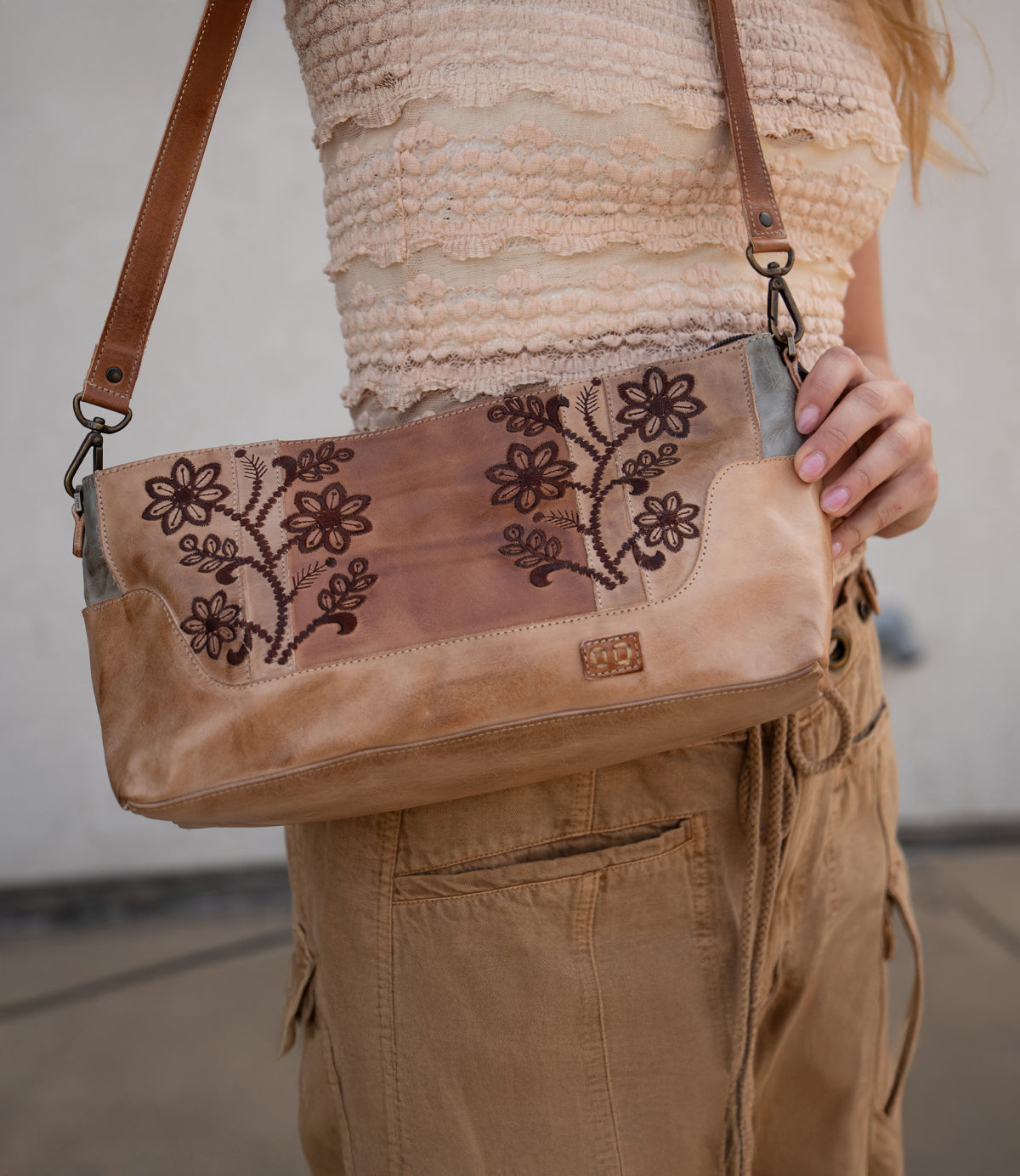 A person in a textured beige top and tan pants holds the Bed Stu Hyperbolic, a handcrafted leather shoulder bag with floral embroidery and a brown strap, adding a chic, bohemian touch.