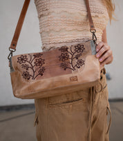 A person in a textured beige top and tan pants holds the Bed Stu Hyperbolic, a handcrafted leather shoulder bag with floral embroidery and a brown strap, adding a chic, bohemian touch.
