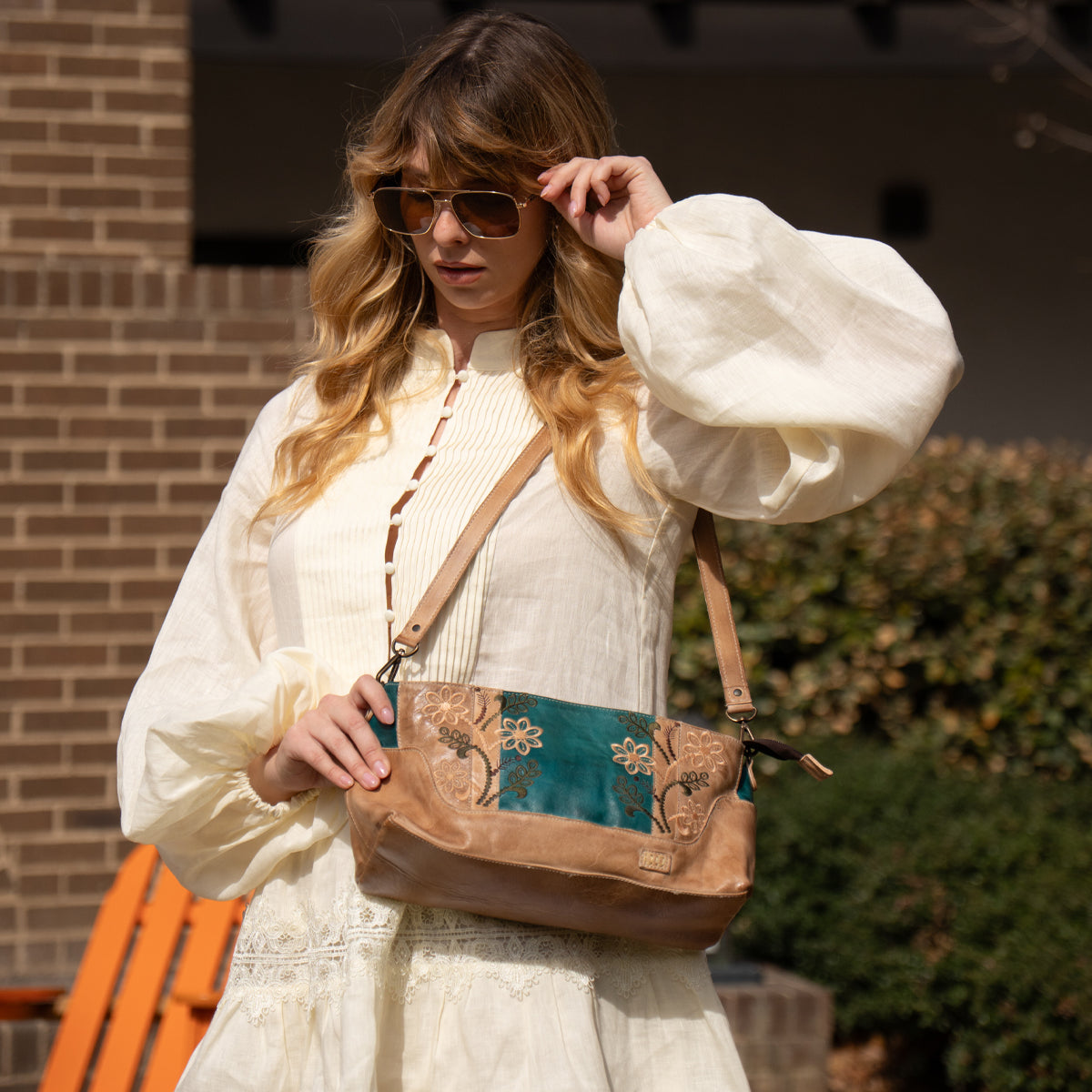 A woman in a cream-colored dress and sunglasses stands outdoors, holding the Bed Stu Hyperbolic handcrafted brown and teal floral leather shoulder bag.