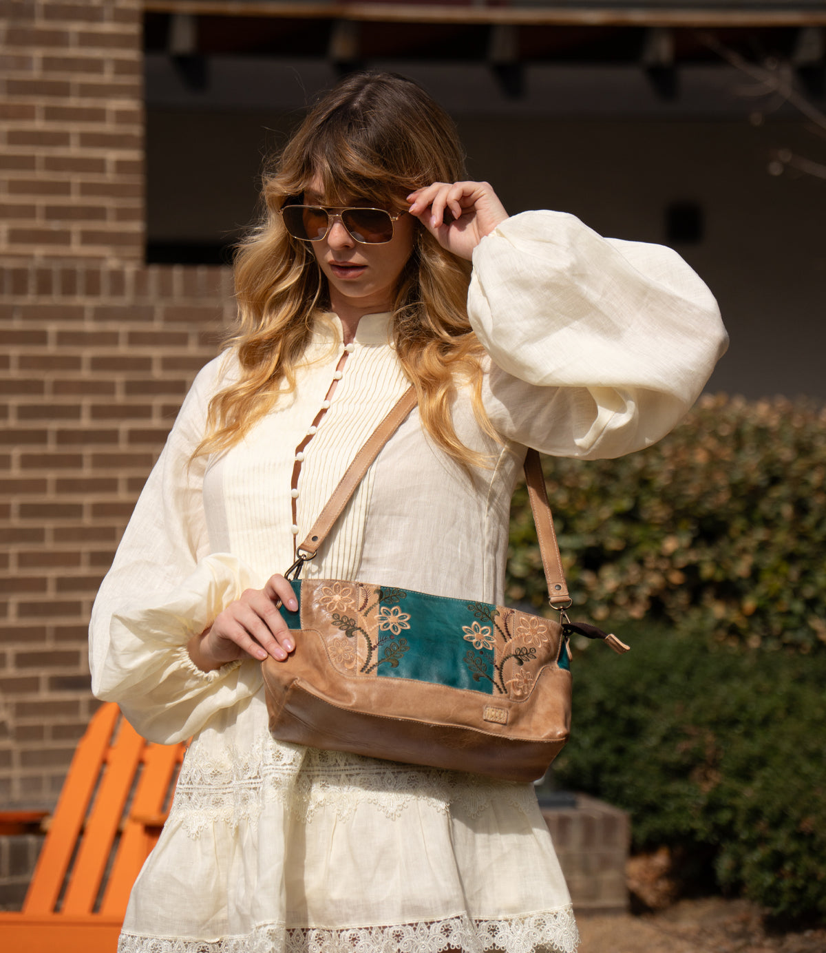 A woman in a cream-colored dress and sunglasses stands outdoors, holding the Bed Stu Hyperbolic handcrafted brown and teal floral leather shoulder bag.