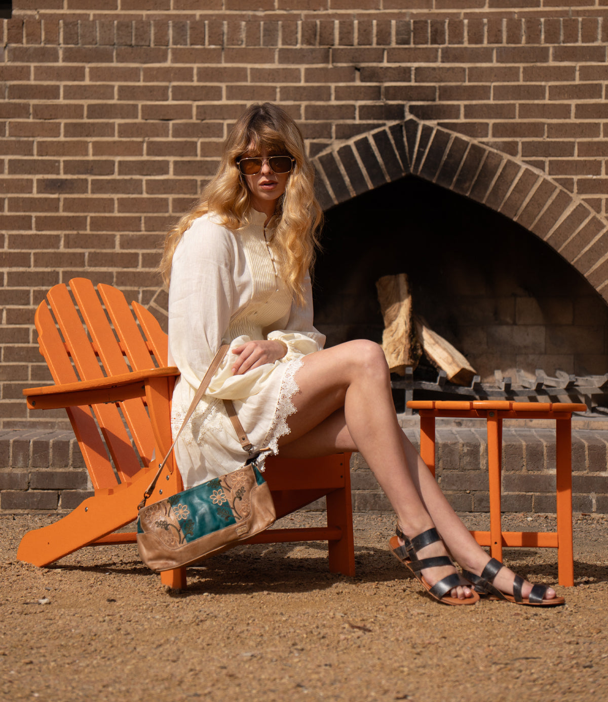 A woman in a white dress and sandals sits on an orange chair by a brick outdoor fireplace, with the Bed Stu Hyperbolic handcrafted leather shoulder bag on her lap and sunglasses on.