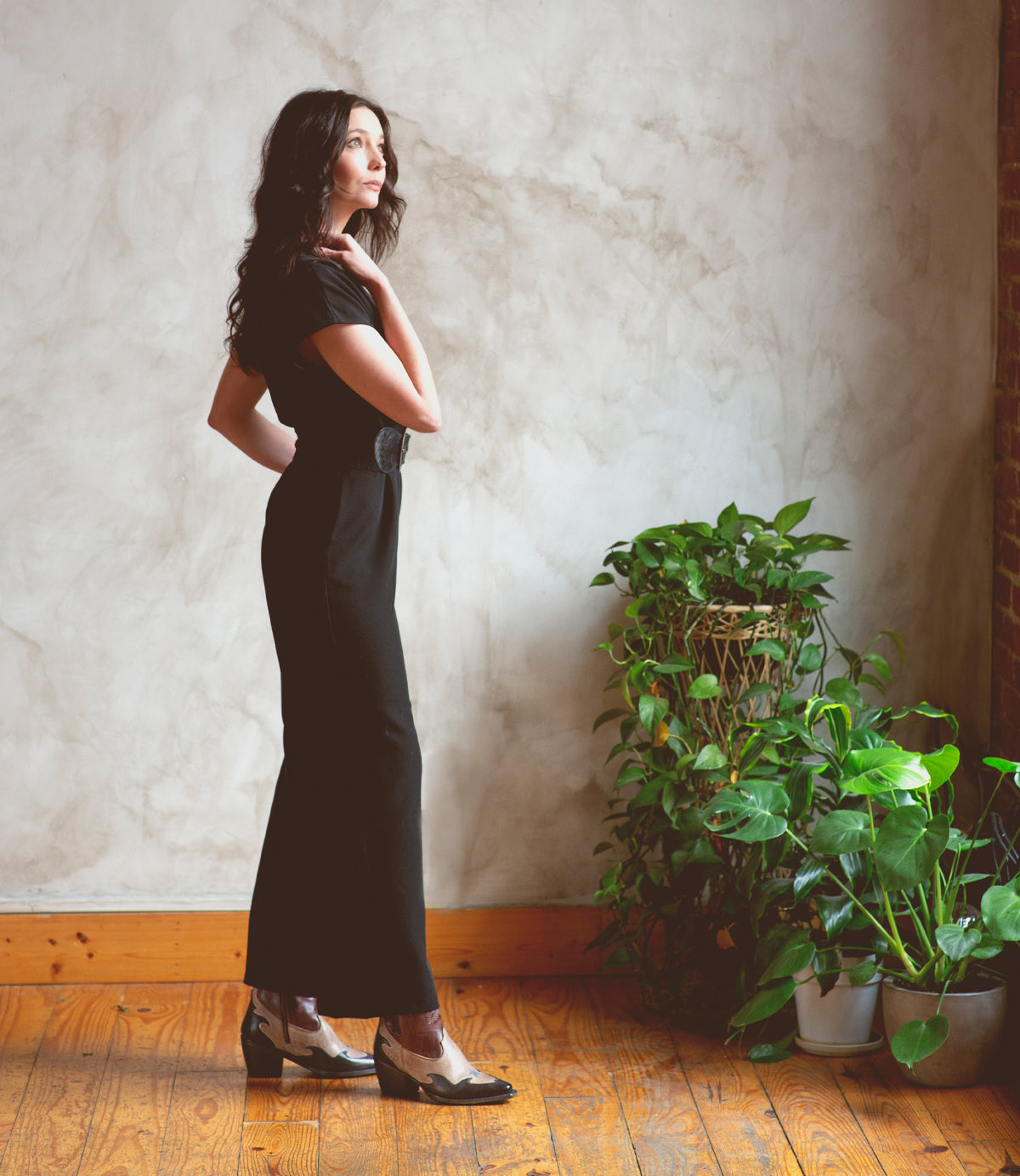 A woman in a black jumpsuit, wearing Bed Stu Hyperspeed boots, standing in front of a potted plant.