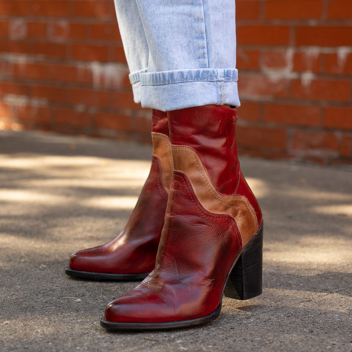 A person wearing light blue cuffed jeans and Bed Stu's Iazu red-brown handcrafted leather boots with a stacked heel stands on a paved sidewalk in front of a red brick wall.