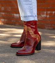 A person wearing light blue cuffed jeans and Bed Stu's Iazu red-brown handcrafted leather boots with a stacked heel stands on a paved sidewalk in front of a red brick wall.