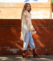 A woman stands sideways by a red brick wall, wearing a sheer white blouse, light blue jeans, and Bed Stu Iazu handcrafted leather boots with a square toe and stacked heel.