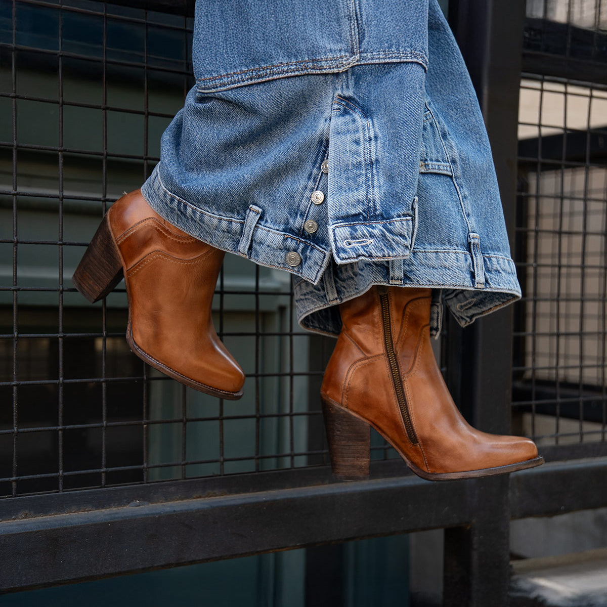 A person wearing wide-leg blue jeans and Bed Stu's Iazu handcrafted brown leather boots with a stacked heel stands on a black metal railing in front of a wire fence.