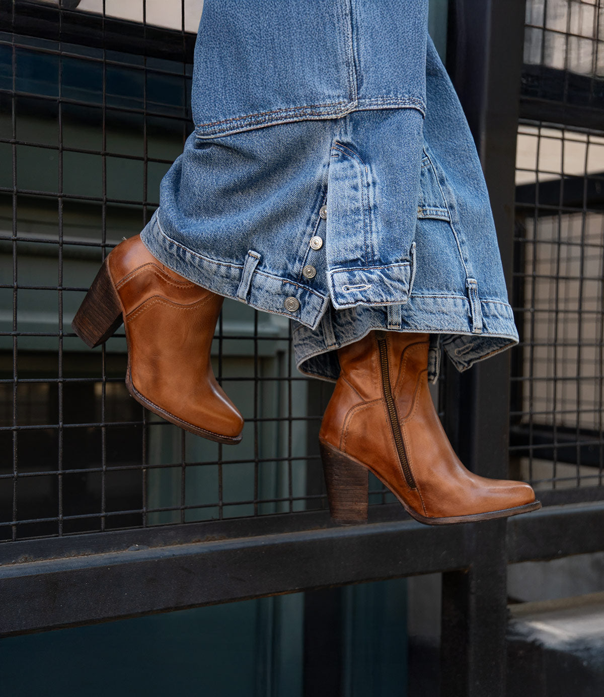 A person wearing wide-leg blue jeans and Bed Stu's Iazu handcrafted brown leather boots with a stacked heel stands on a black metal railing in front of a wire fence.