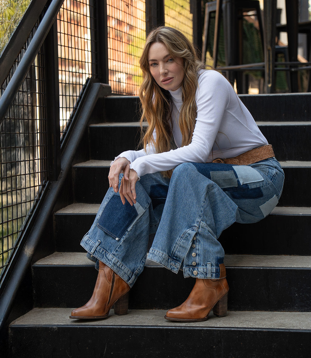 A woman with long hair sits on outdoor stairs, wearing a white long-sleeve top, patchwork denim jeans, a brown belt, and Bed Stu’s handcrafted Iazu leather boots with a stacked heel.