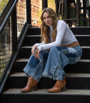 A woman with long hair sits on outdoor stairs, wearing a white long-sleeve top, patchwork denim jeans, a brown belt, and Bed Stu’s handcrafted Iazu leather boots with a stacked heel.