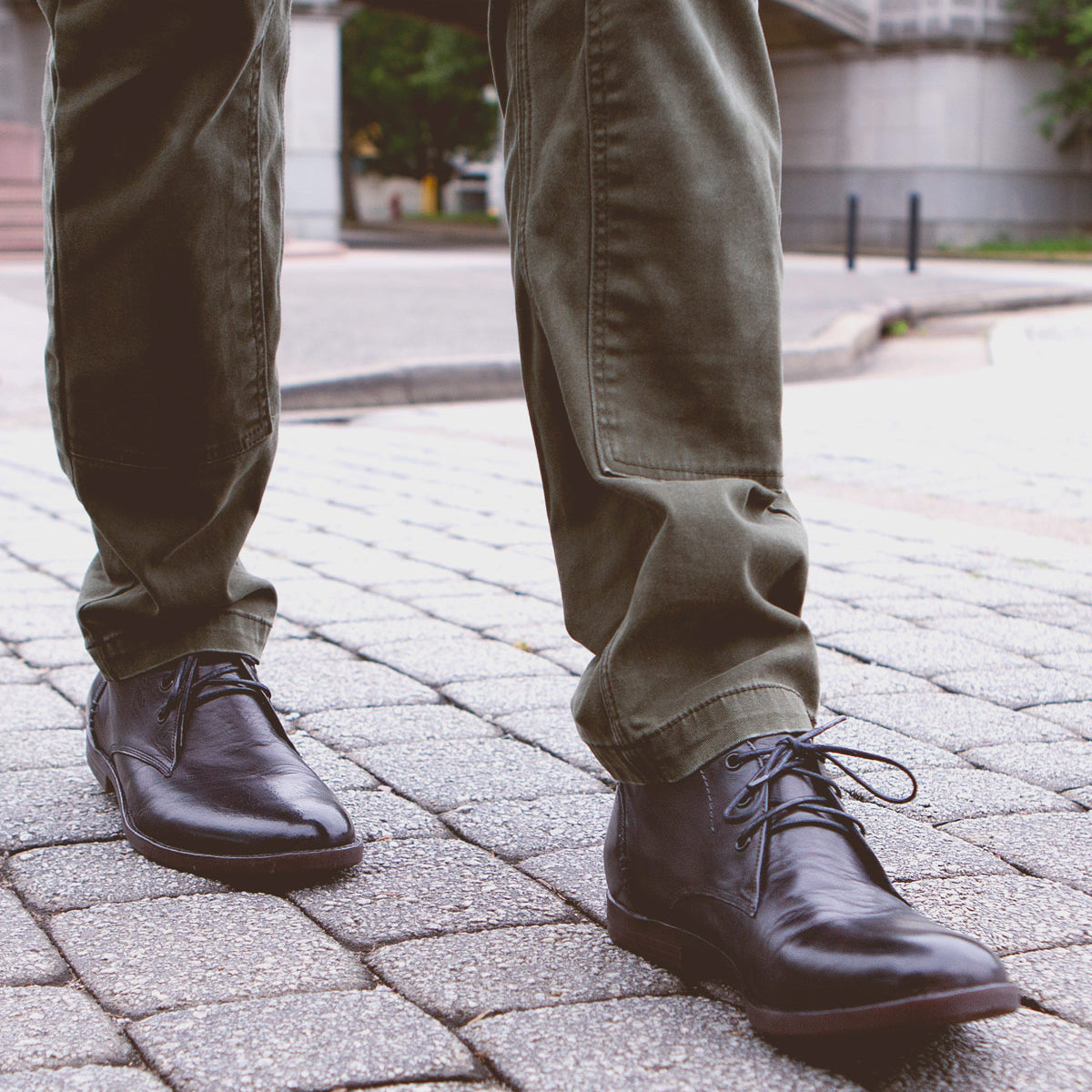 Close-up view of a person wearing green pants and black leather chukka boots with a newly developed outsole, Illiad by Bed Stu, standing on a cobblestone street.