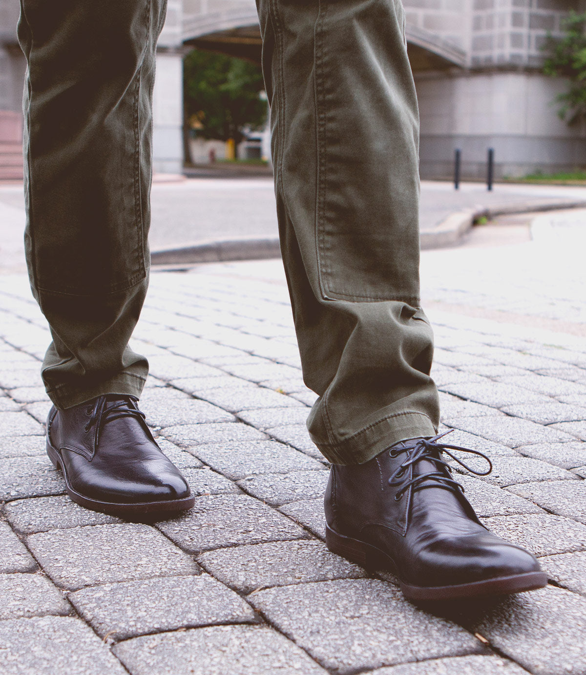 Close-up view of a person wearing green pants and black leather chukka boots with a newly developed outsole, Illiad by Bed Stu, standing on a cobblestone street.