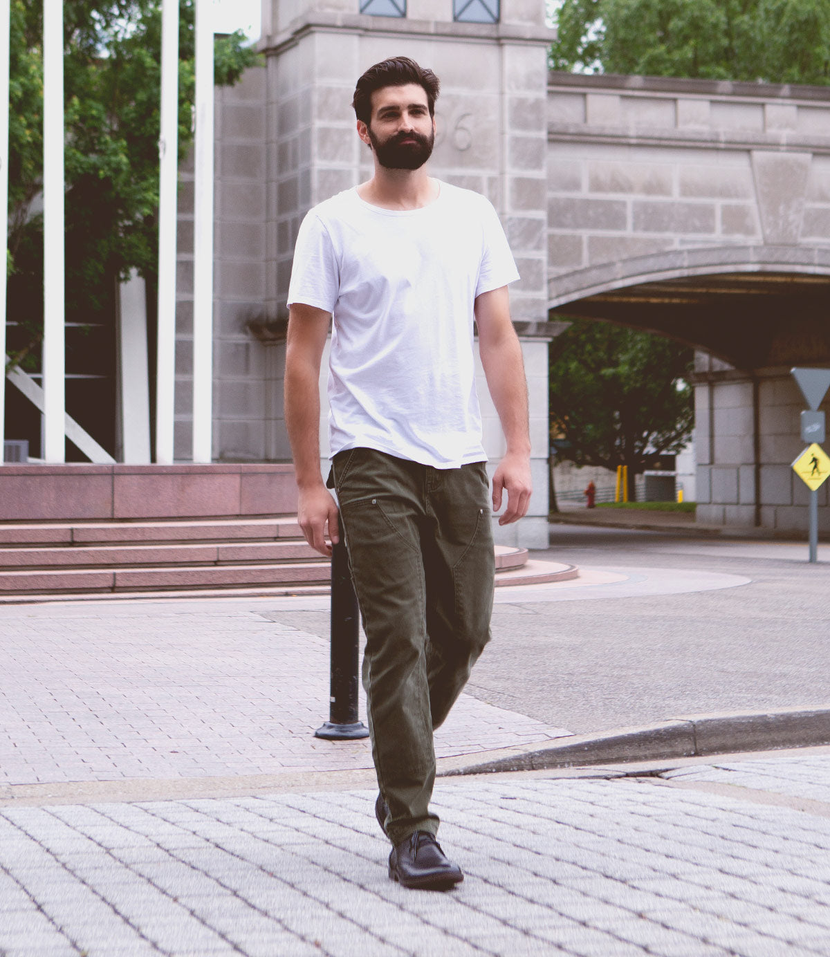 A man with a beard, wearing a white t-shirt, green pants, and leather chukka boots with a newly developed Illiad outsole by Bed Stu, walks on a paved path near a large stone building with an archway.