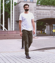 A man with a beard, wearing a white t-shirt, green pants, and leather chukka boots with a newly developed Illiad outsole by Bed Stu, walks on a paved path near a large stone building with an archway.