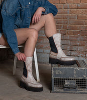 A person sits on a chair in Bed Stu's Illicit denim jacket and chunky platform ankle boots, exuding retro flair against a brick wall and metal crate backdrop.