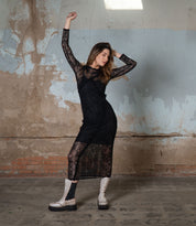 In a textured room, a woman exudes nostalgic fashion in a long black lace dress and completes her retro flair with Bed Stu's chunky white "Illicit" platform ankle boots, posing elegantly with raised arms.