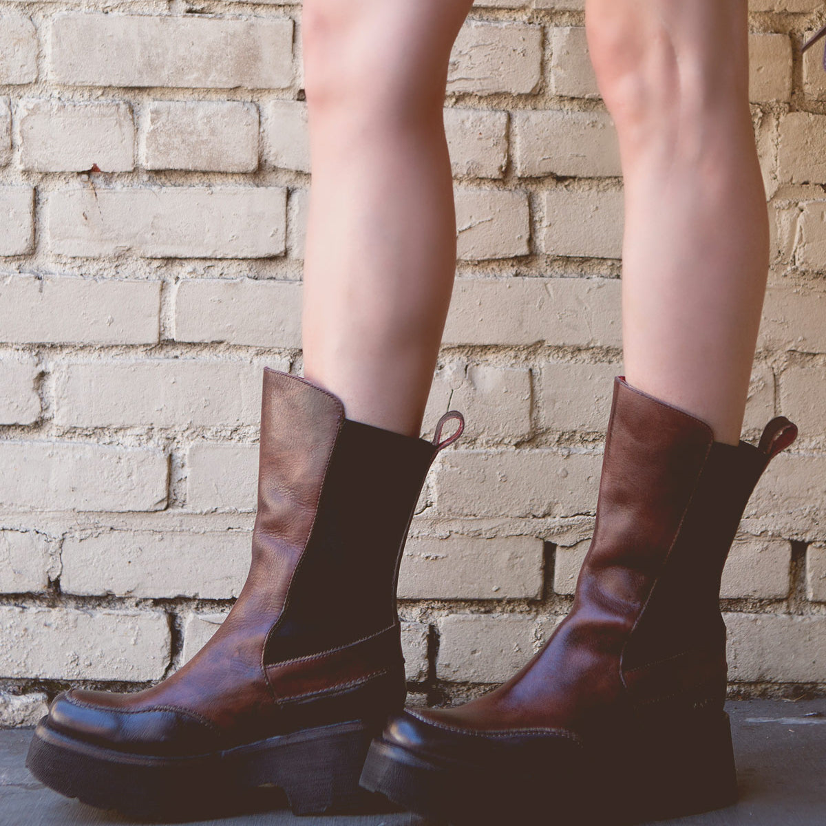 A person wearing Bed Stu's Illicit, a pair of brown leather boots with a touch of retro flair, stands on a concrete surface in front of a brick wall.
