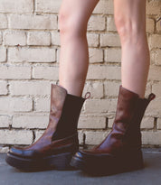 A person wearing Bed Stu's Illicit, a pair of brown leather boots with a touch of retro flair, stands on a concrete surface in front of a brick wall.