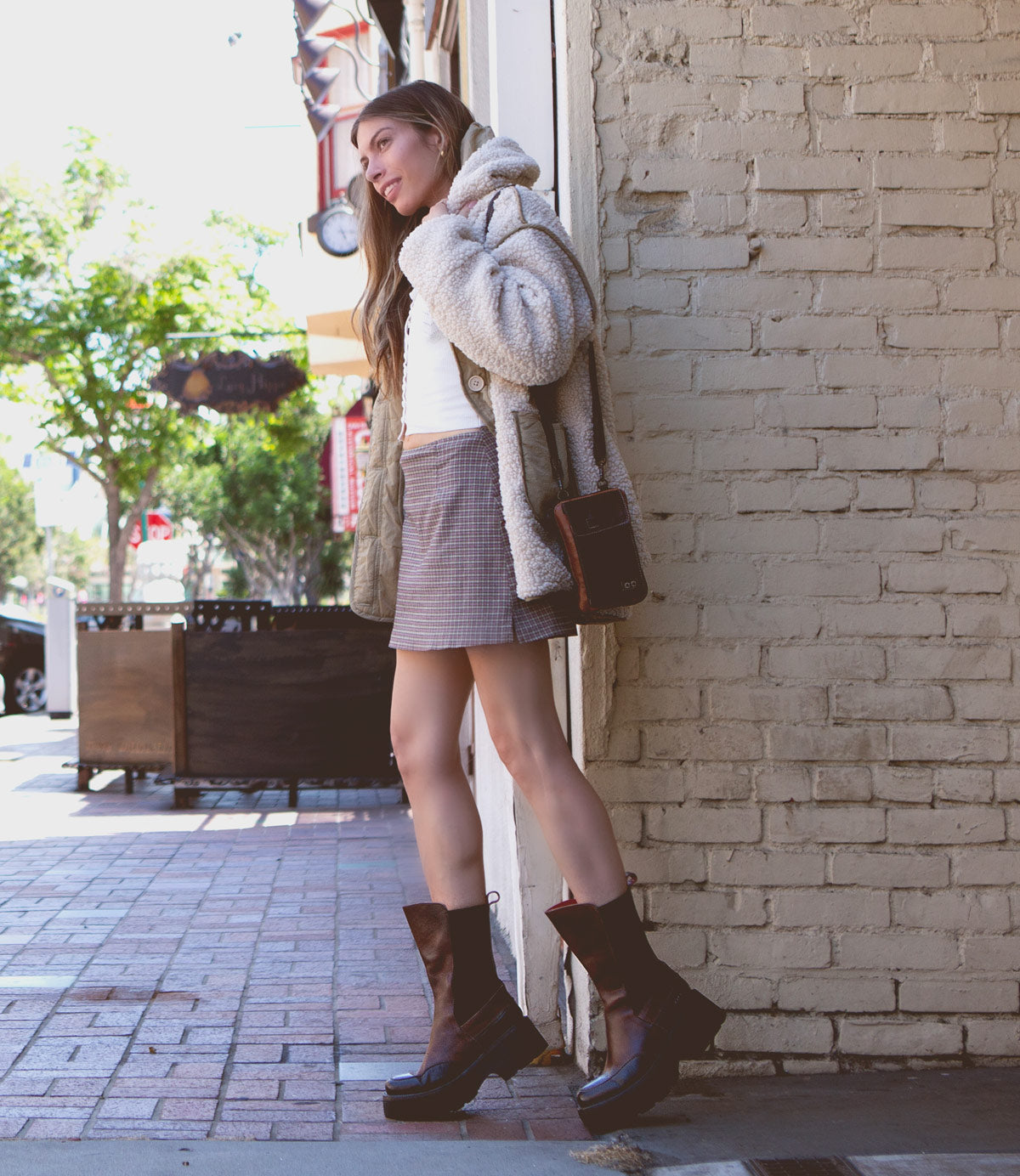 A person stands against a brick wall, wearing the "Illicit" jacket from Bed Stu paired with a skirt and chunky platform ankle boots, exuding a retro flair. In the background, a sidewalk and tree complete this nostalgic fashion moment.