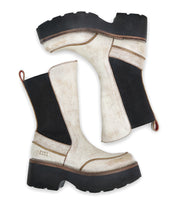 The "Illicit" boots from Bed Stu, featuring a beige and black design with chunky soles and red pull tabs, exude retro flair against a white background.