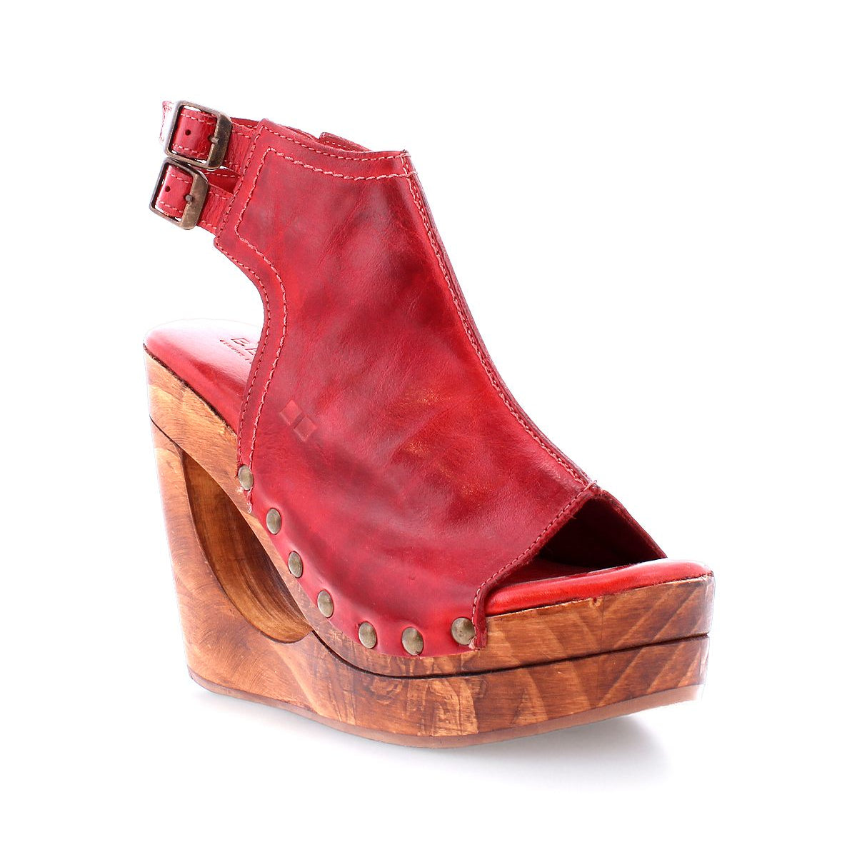 The Imelda by Bed Stu is a red open-toe wedge sandal, featuring a wooden platform and metal studs, complete with an ankle strap adorned with a stylish buckle.