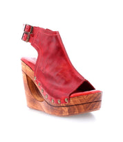 The Imelda by Bed Stu is a red open-toe wedge sandal, featuring a wooden platform and metal studs, complete with an ankle strap adorned with a stylish buckle.