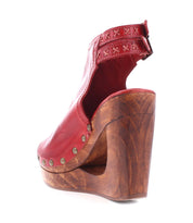 The Imelda by Bed Stu is a red leather sandal featuring an open-toe design and ankle strap, complemented by a wooden wedge heel embellished with metal studs along the base.