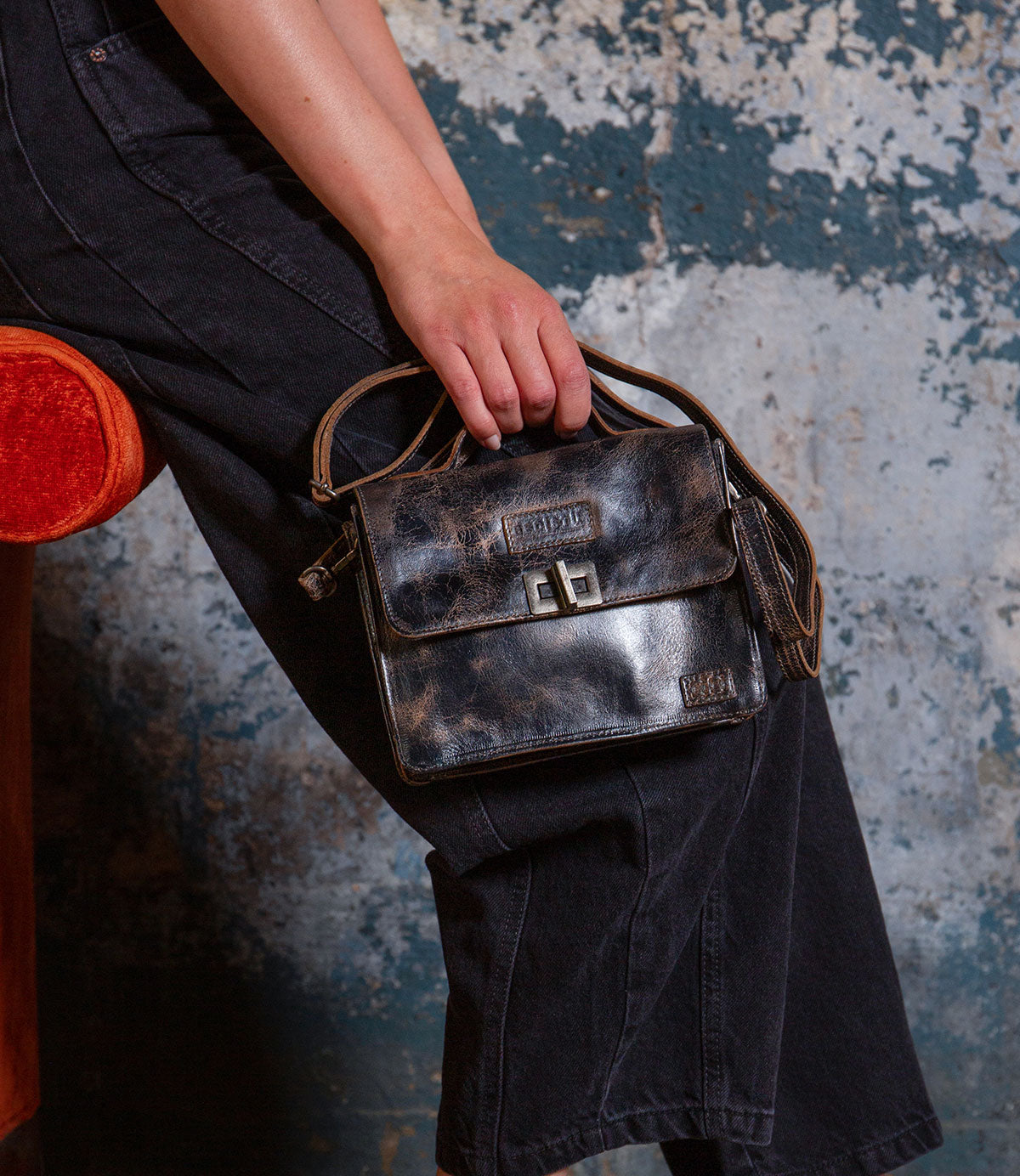 A person in black pants sits on an orange chair, holding the Bed Stu Improv—a small, worn black leather satchel bag with a metal clasp and top handle.