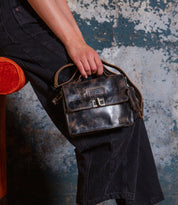 A person in black pants sits on an orange chair, holding the Bed Stu Improv—a small, worn black leather satchel bag with a metal clasp and top handle.