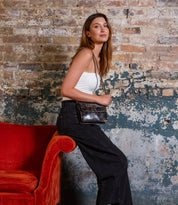 A woman in a white tank top and black pants sits on a red chair’s arm, wearing the Bed Stu Improv mini purse with an adjustable crossbody strap, set against a distressed brick wall.