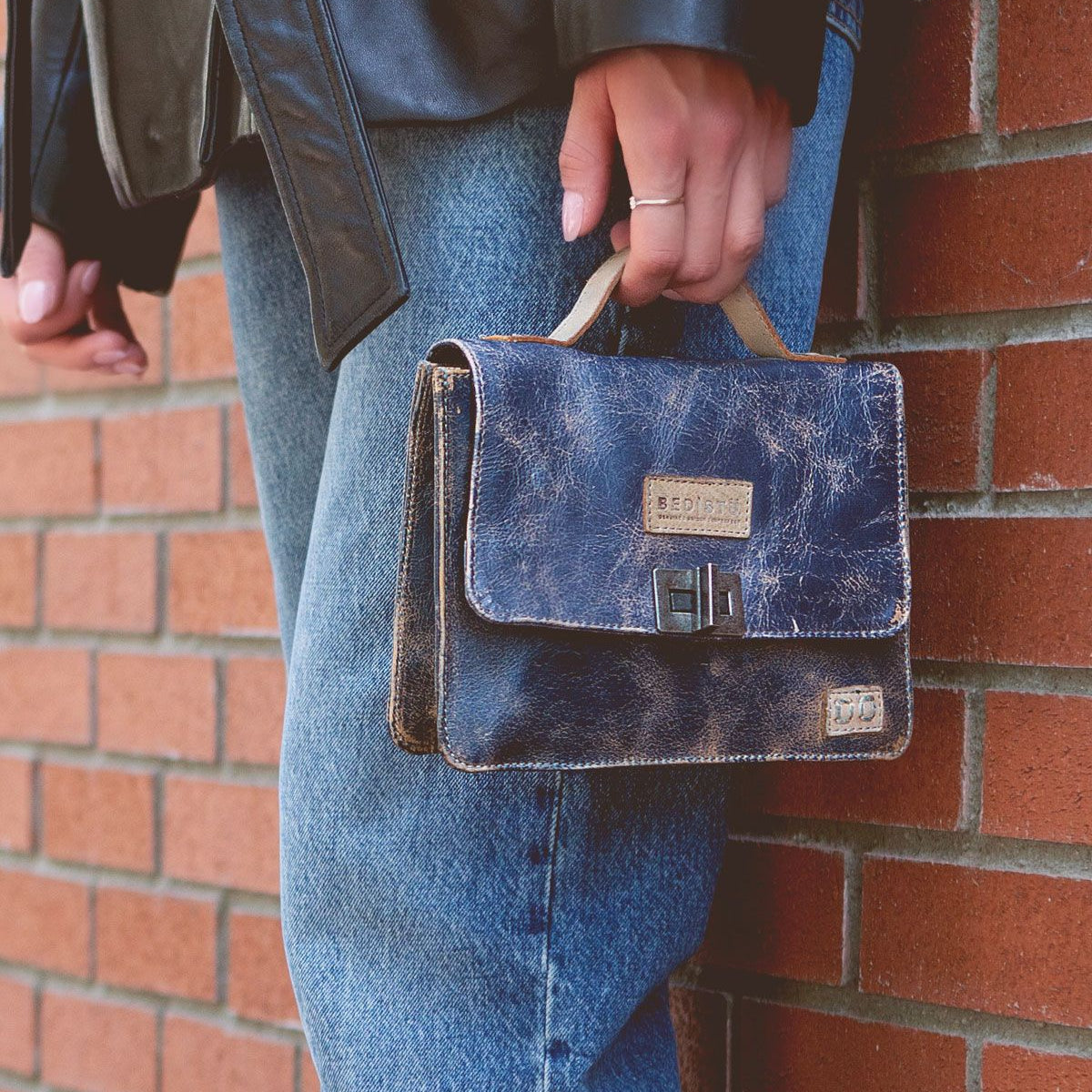 A person in blue jeans and a black jacket holds the Bed Stu Improv, a small, worn leather satchel with a crossbody strap, against a brick wall, blending fashion and functionality seamlessly.