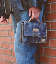 A person in blue jeans and a black jacket holds the Bed Stu Improv, a small, worn leather satchel with a crossbody strap, against a brick wall, blending fashion and functionality seamlessly.