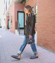 A woman in a black coat and rolled-up jeans walks on a brick sidewalk near a brick building, carrying the stylish Improv mini purse by Bed Stu.