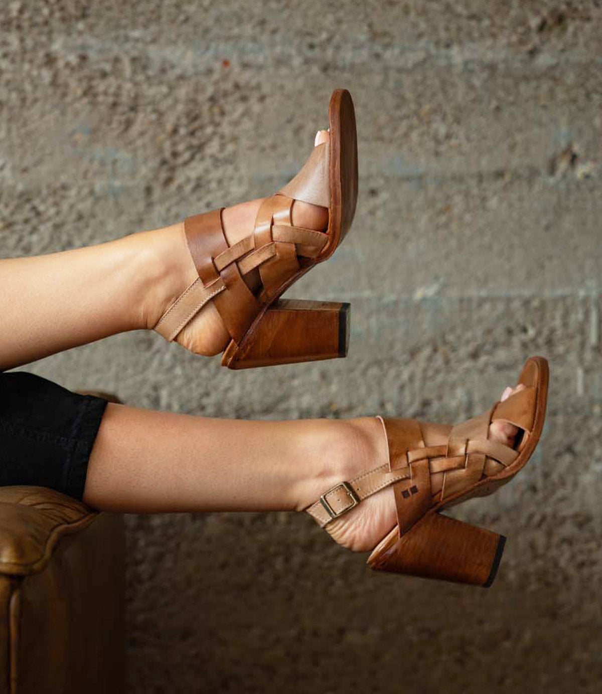 A person wears the Bed|Stü Intersection tan woven leather heeled sandals, featuring an open-toe design and adjustable ankle strap, while resting their feet on a brown chair against a textured gray wall.