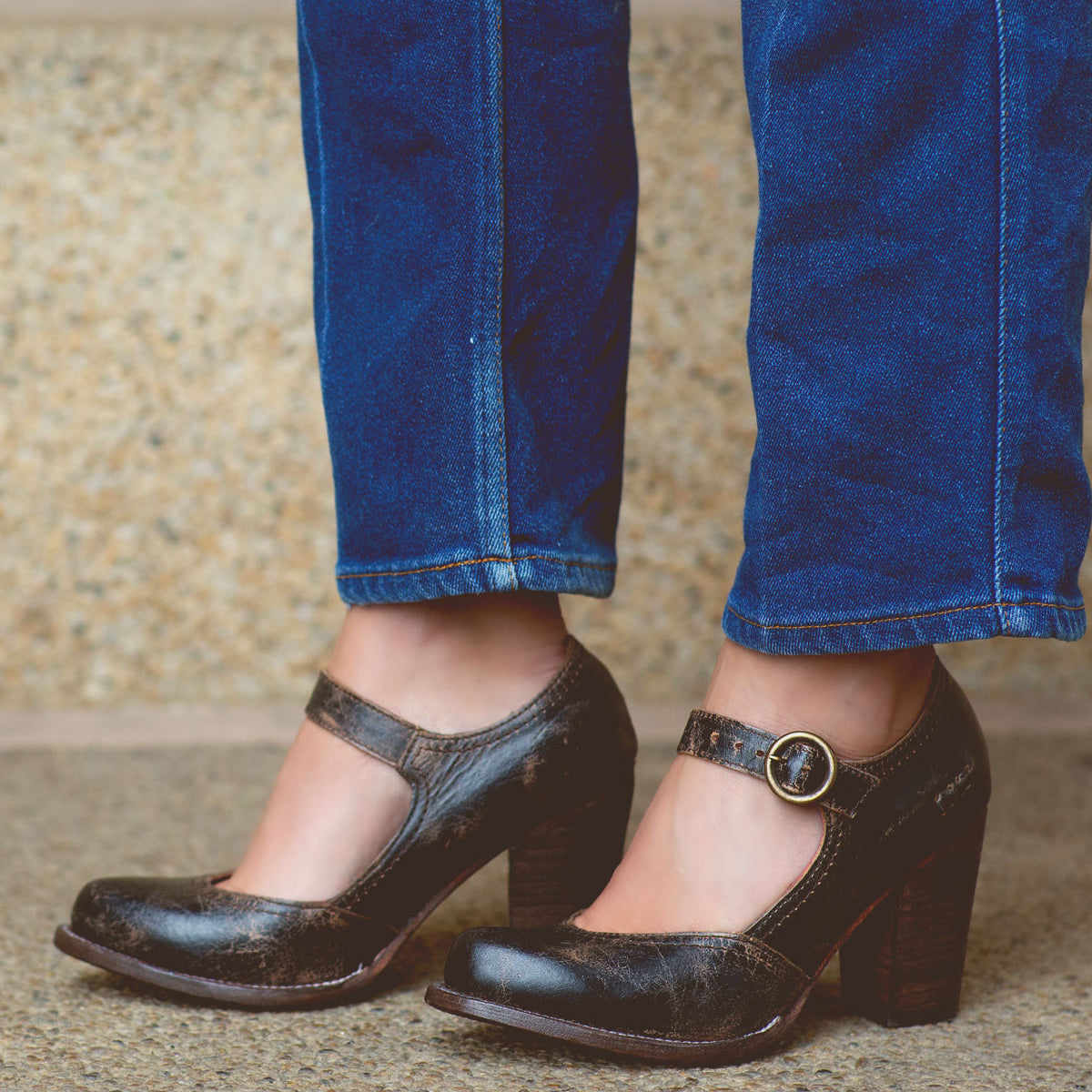 A person wearing dark brown Bed Stu Ishtar pumps with an adjustable strap and blue jeans stands on a concrete surface with stone steps in the background.