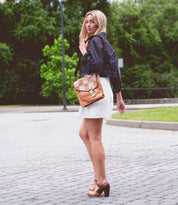 A blonde woman wearing a denim jacket and white dress stands on a cobblestone road with trees in the background, carrying an Ishtar by Bed Stu, a tan shoulder bag with an adjustable strap, and wearing brown block heels.
