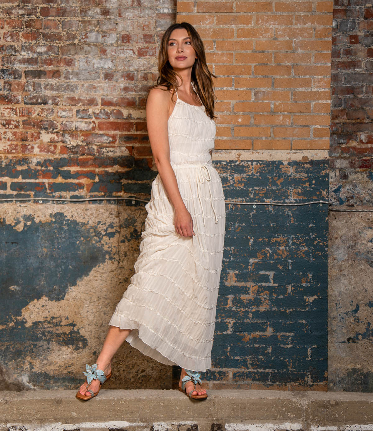 A woman wearing Bed Stu's Isobel sleeveless white dress and blue braided leather sandals stands before a weathered brick-and-painted wall, gazing slightly to the side.