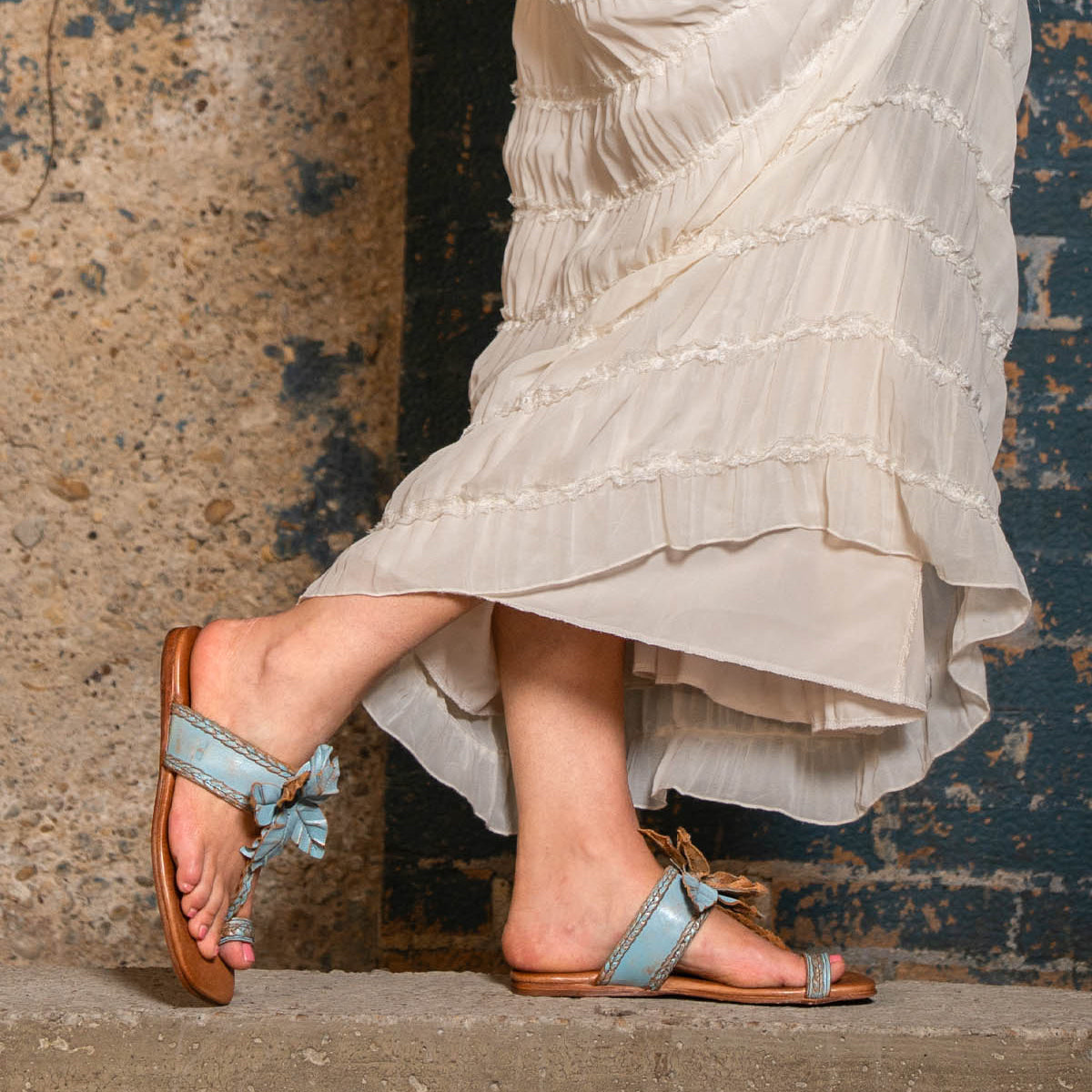 Wearing Bed Stu’s Isobel light blue braided leather sandals, a person in a long cream ruffled skirt stands on a concrete step against a textured wall.