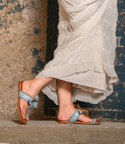 Wearing Bed Stu’s Isobel light blue braided leather sandals, a person in a long cream ruffled skirt stands on a concrete step against a textured wall.
