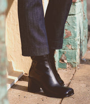 A person stands confidently on the sidewalk dressed in dark jeans and sporting the sleek Isodora ankle boots by Bed Stu. Made from handcrafted Italian leather, these black boots feature an asymmetrical square toe and block heels, adding a touch of avant-garde elegance to their ensemble.