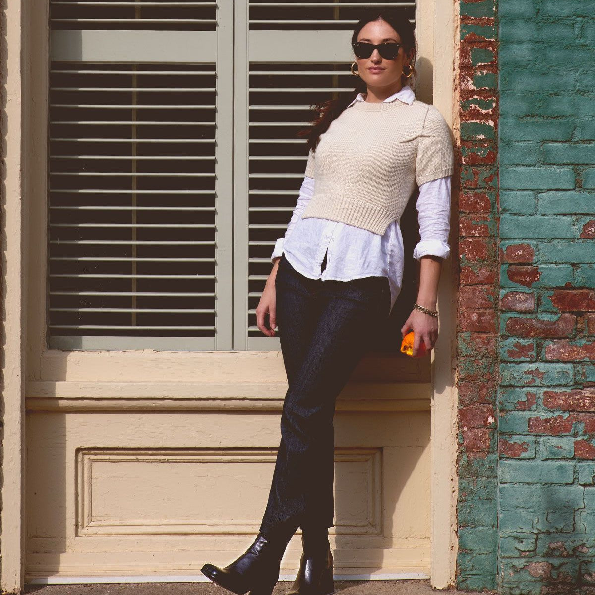A person in a white shirt and sweater vest stands against a building wall, showcasing handcrafted Bed Stu leather boots named Isodora while holding a small orange object.