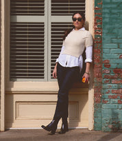 A person in a white shirt and sweater vest stands against a building wall, showcasing handcrafted Bed Stu leather boots named Isodora while holding a small orange object.