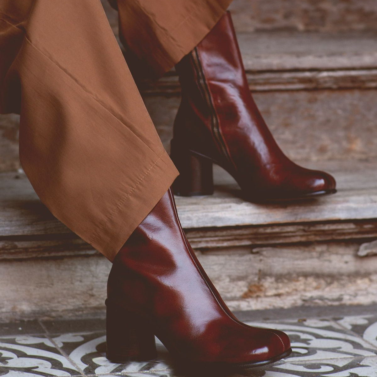 A person wearing the Isodora boots by Bed Stu, made from dark brown handcrafted Italian leather with an asymmetrical square toe, pairs them with brown pants while standing on patterned tile steps.