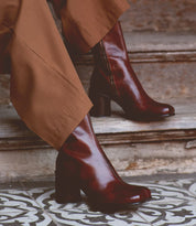 A person wearing the Isodora boots by Bed Stu, made from dark brown handcrafted Italian leather with an asymmetrical square toe, pairs them with brown pants while standing on patterned tile steps.