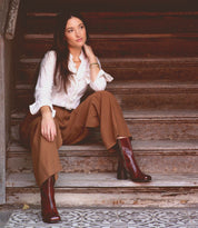 A woman in a white blouse and brown pants sits on wooden stairs, gazing thoughtfully. She wears the Isodora by Bed Stu, handcrafted Italian leather boots featuring an asymmetrical square toe.