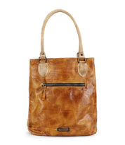 The Italic by Bed Stu is a tan leather tote bag with a timeless design, featuring two handles, a front zipper pocket, and a small logo tag at the bottom. It offers a spacious interior.