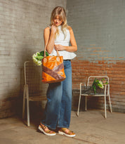 A woman with blond hair in a white top and jeans holds an Italic leather tote by Bed Stu, filled with flowers. She stands in a spacious room featuring a brick wall and two white chairs decorated with plants.