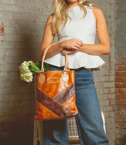 A person in a sleeveless white top and blue jeans holds the elegant Bed Stu Italic leather tote bag with an X-pattern. Flowers peek out of its spacious interior, complementing the stylish ensemble, while a weathered brick wall forms the backdrop.