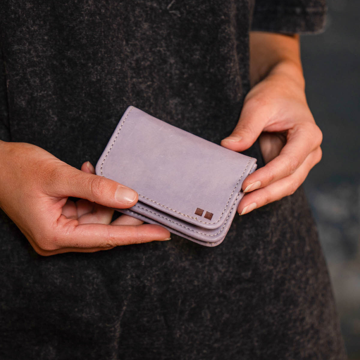 Person holding the Jeor card holder by Bed Stu—a small, light purple rustic leather accessory with stitched edges, credit card slots, and a metal square detail on the front.