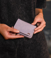 Person holding the Jeor card holder by Bed Stu—a small, light purple rustic leather accessory with stitched edges, credit card slots, and a metal square detail on the front.